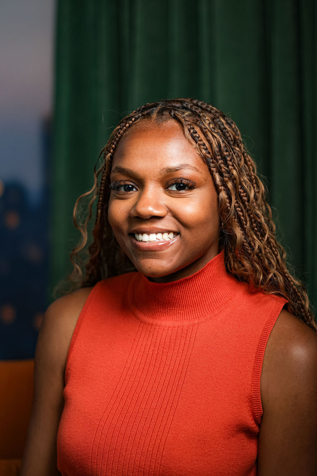 Therapist sitting smiling with an orange shirt, braids, and a green curtain in the background.