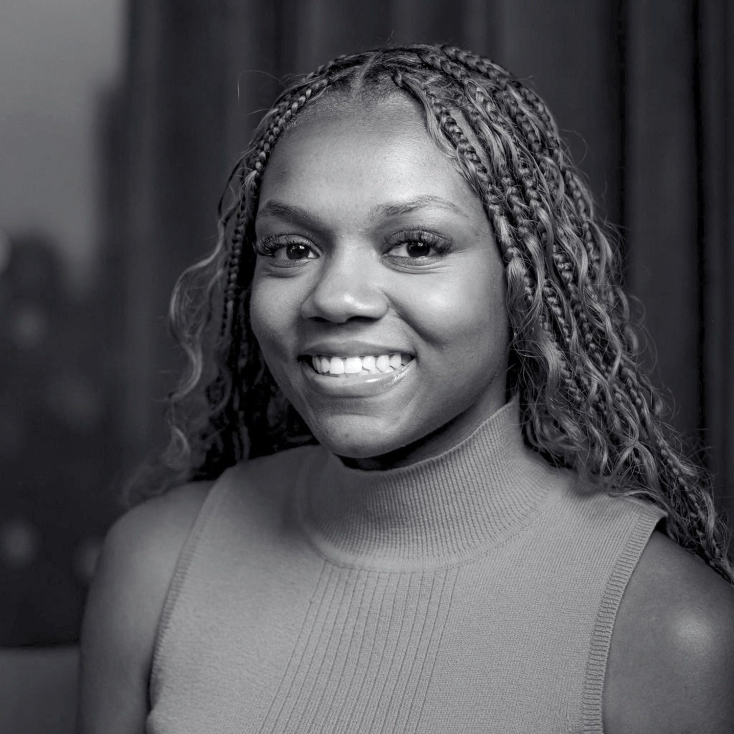 A black and white portrait of a smiling woman with braided hair wearing a ribbed sleeveless turtleneck top.